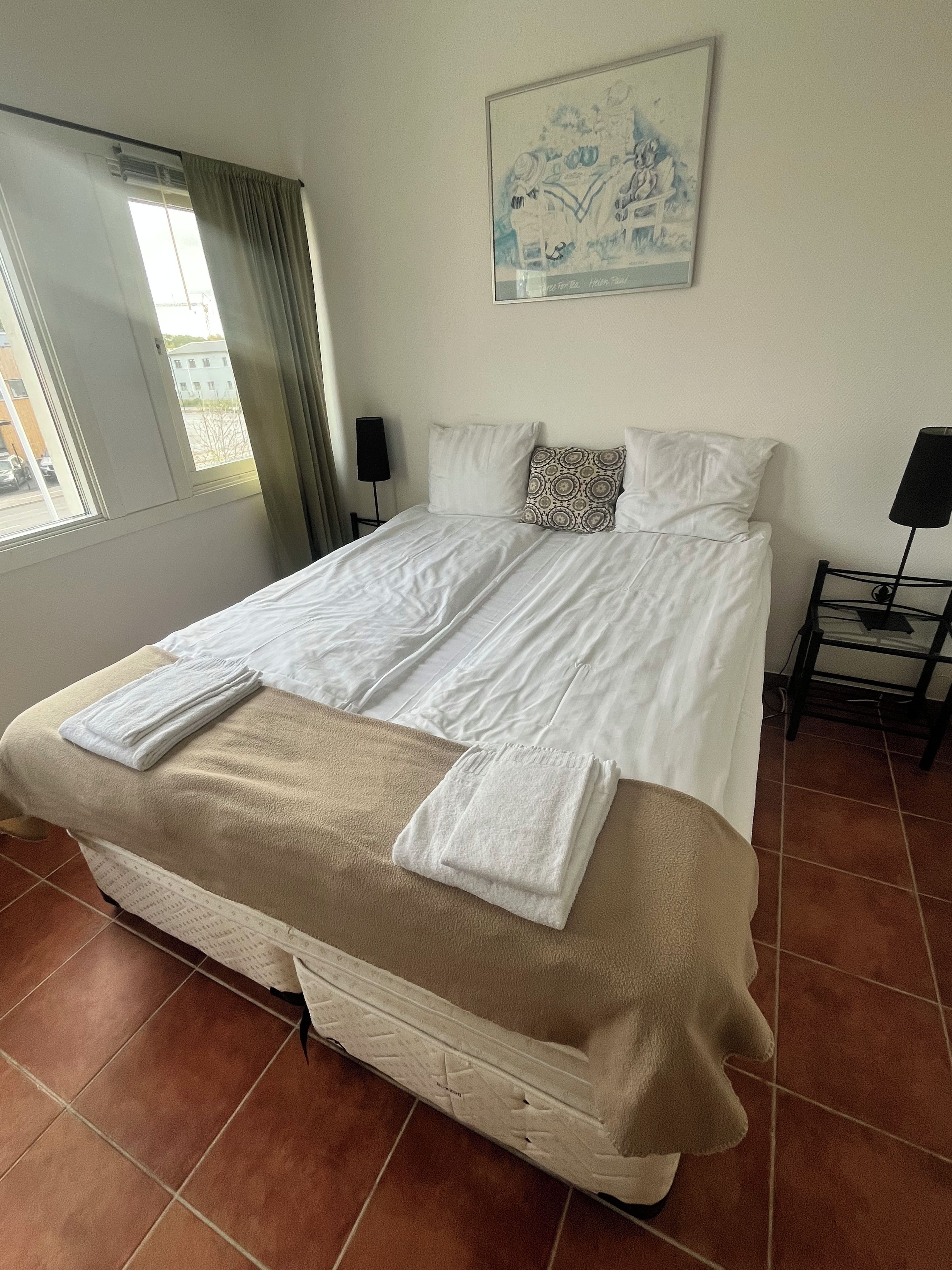 Habitación de hotel con una cama grande, un cuadro en la pared, una ventana con cortinas y piso de baldosas.