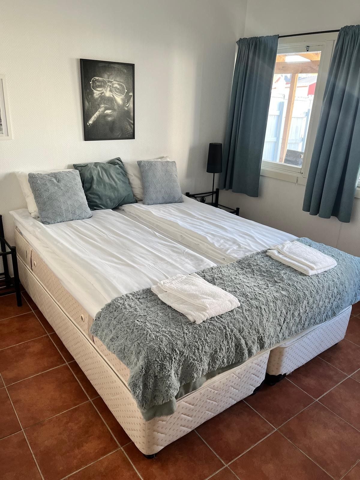 A bedroom with a large bed, a bedside table, a lamp, a window with curtains, and a wall-mounted picture.