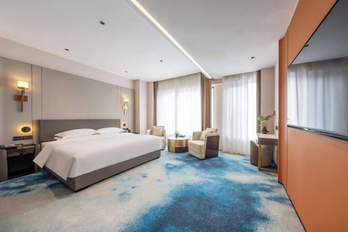 Een moderne hotelkamer met een groot bed, nachtkastjes, een zithoek met een fauteuil en een klein tafeltje, en een groot raam met gordijnen.