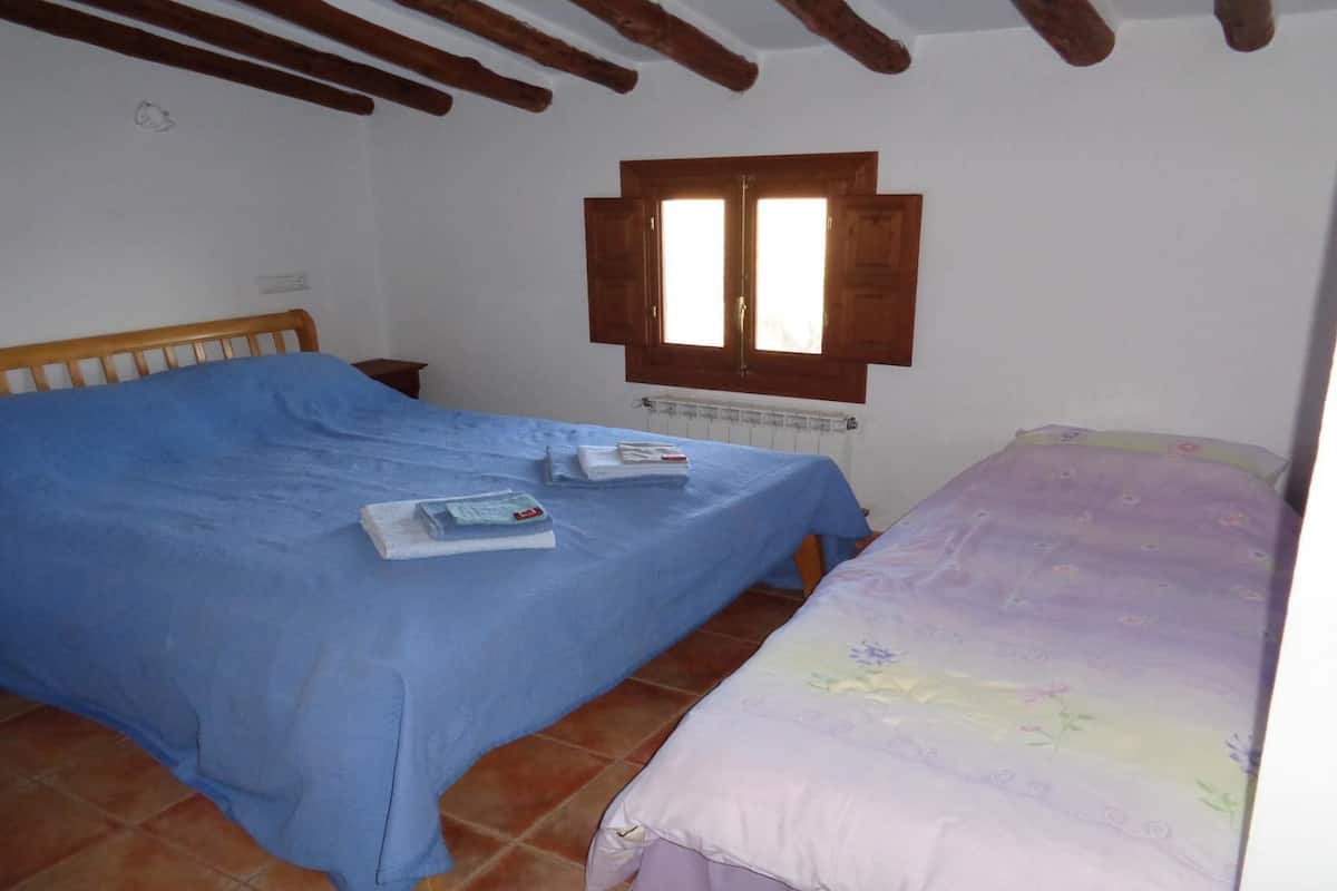 Une chambre avec deux lits, l’un recouvert d’une couvre-lit bleue et l’autre d’une couvre-lit à motifs floraux. Il y a une fenêtre avec des volets en bois, un radiateur en dessous et une poutre en bois au plafond.