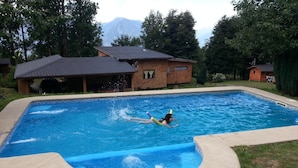 Una piscina con una persona nadando, rodeada de césped y una casa con tejado de baldosas.