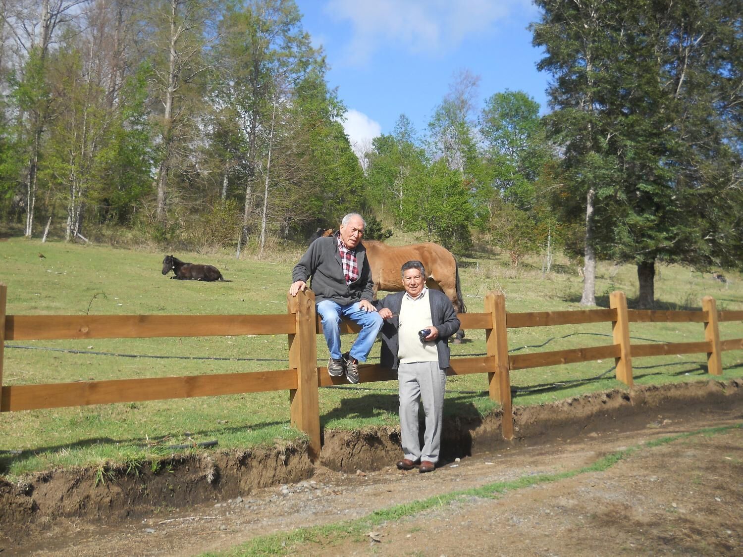 Two people sitting on a wooden fence in a rural setting with a horse in the background.