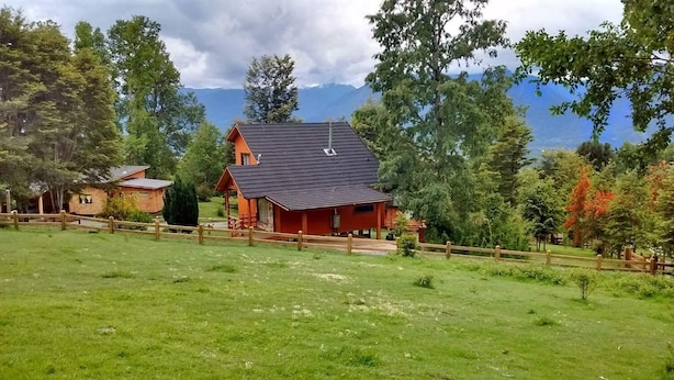 Una cabaña roja con techo inclinado, rodeada por una cerca de madera y abundante vegetación.