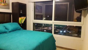 A bedroom with a bed, a window with a city view, and a bookshelf. - Departamento Gran Marina (La Serena)