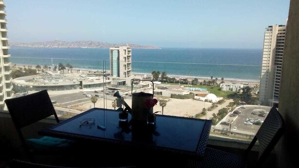 A balcony with a table and chairs overlooking a coastal cityscape with a beach and high-rise buildings. - Departamento Gran Marina (La Serena)