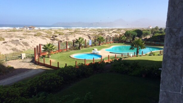 A view of a property with two swimming pools surrounded by a fence, a grassy area, and a view of the ocean. - Depto 203 Fragata Condominio Playa (La Serena)