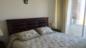 A bed with a floral-patterned quilt, a wooden headboard, two bedside lamps, and a window with curtains. - Depto 203 Fragata Condominio Playa (La Serena)