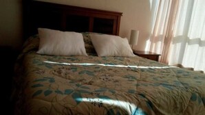 A bed with a floral bedspread, two pillows, a wooden headboard, and a nightstand with a lamp. - Piscina Cristalina Coral (La Serena)