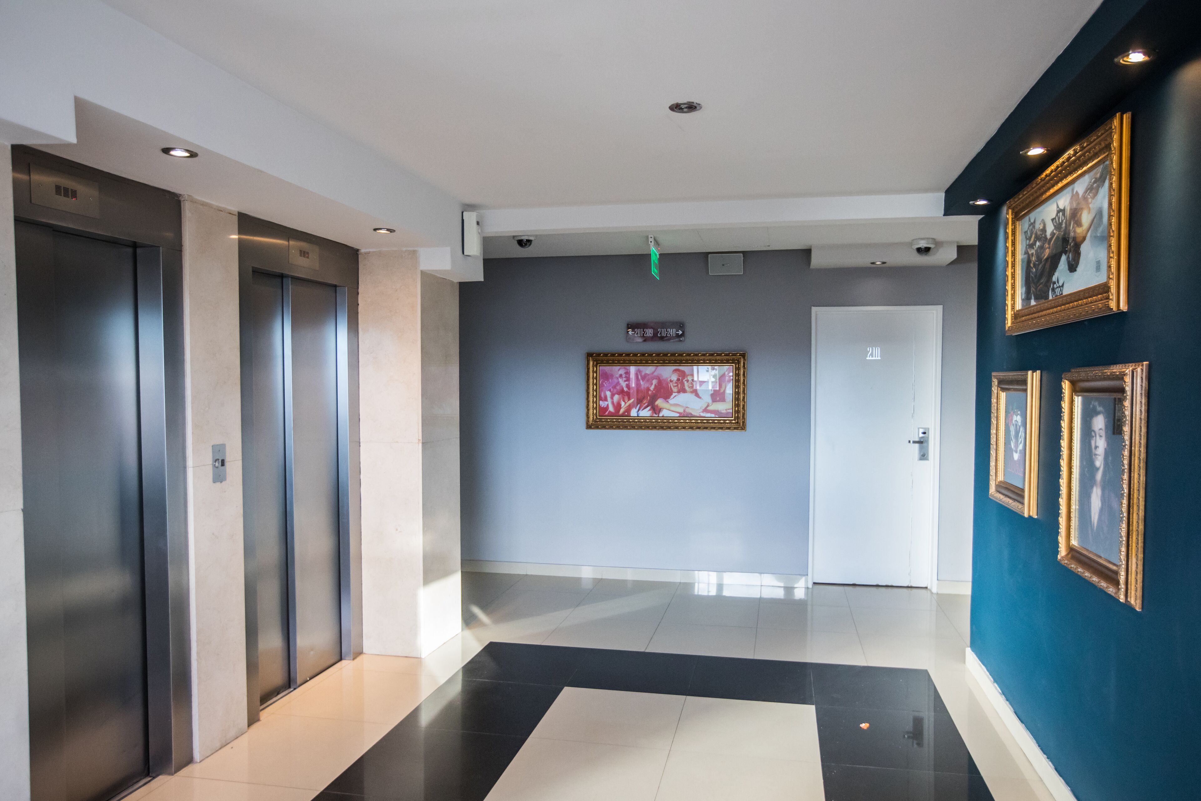A modern hallway with elevators, a door labeled "211", and framed artwork on the walls.