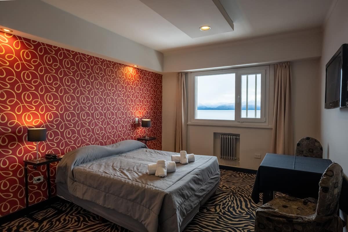 A hotel room with a bed, a desk, a chair, a window with a view, and patterned wallpaper.