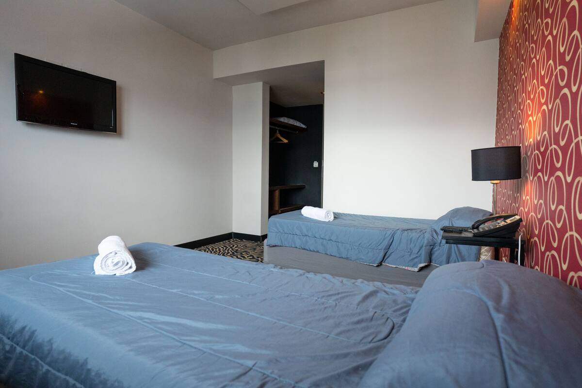 A hotel room with two beds, a flat-screen TV mounted on the wall, a desk with a lamp, and a patterned wall.
