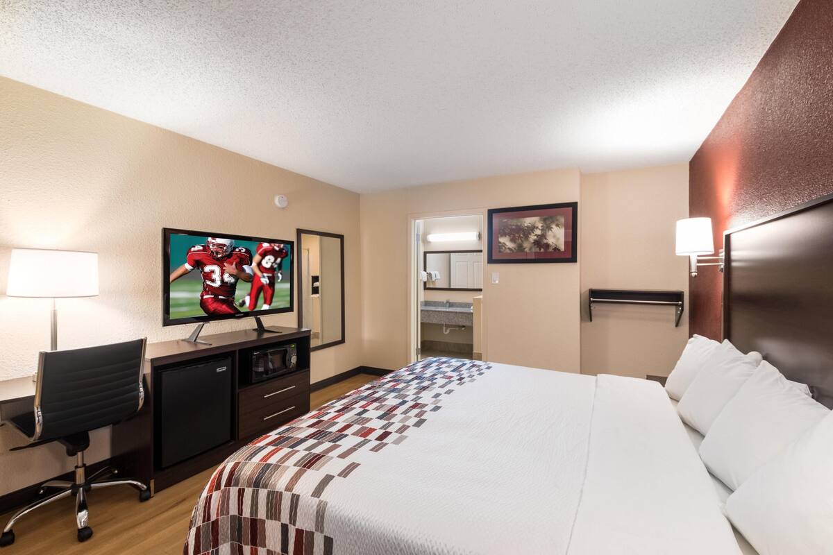 A hotel room with a bed, a desk with a computer, a TV, a mirror, and a bathroom visible through an open door.