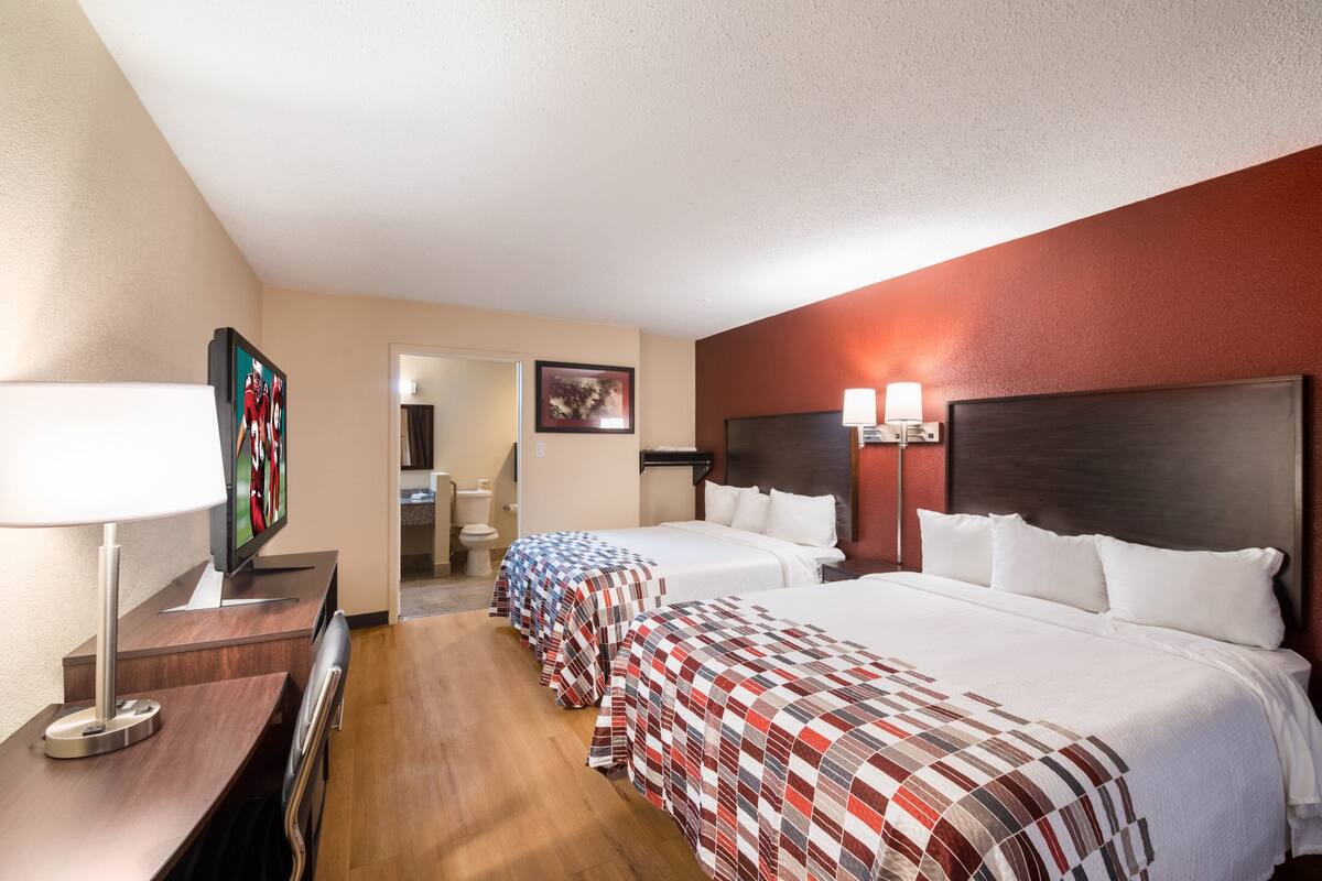 A hotel room with two beds, a desk with a lamp, a TV, and a bathroom visible in the background.