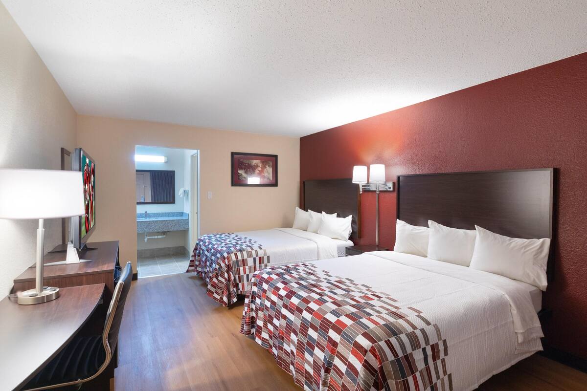 A hotel room with two beds, a desk, a chair, a lamp, and a painting on the wall.