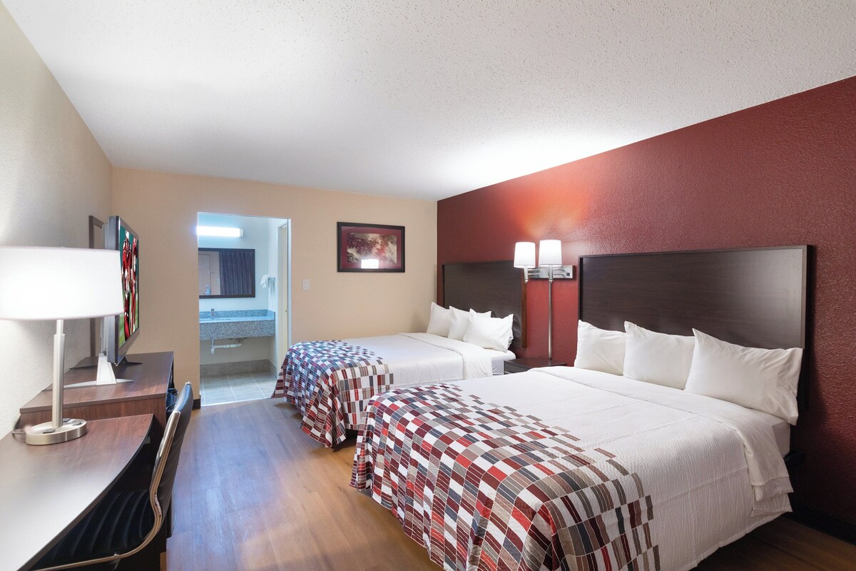 A hotel room with two beds, a desk with a lamp, a TV, and a bathroom visible through an open door.