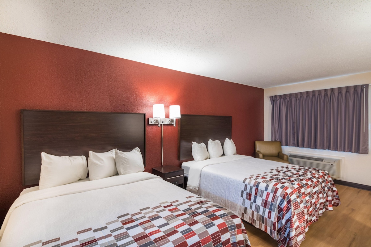 A hotel room with two beds, a nightstand, a lamp, a chair, and a window with curtains.