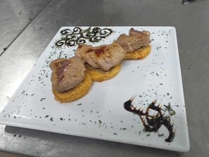 Un plato cuadrado y blanco con tres porciones de carne a la parrilla sobre una base de pan amarillo, adornado con hierbas y salsa oscura.