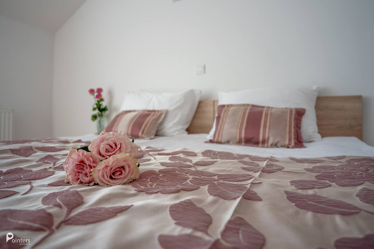 Cama bem arrumada com colcha florida, um buquê de rosas cor-de-rosa e almofadas listradas.