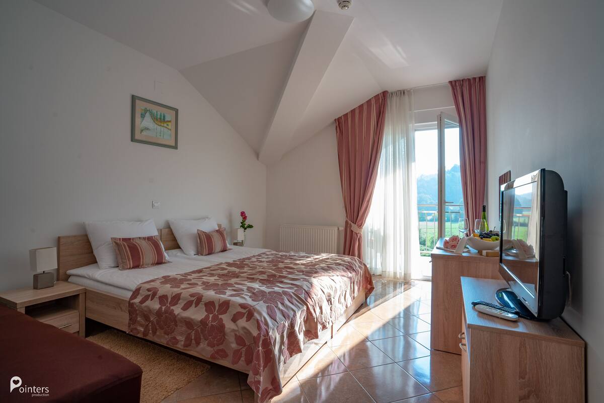 Quarto com cama, escrivaninha com televisão, janela com cortinas e um quadro na parede.