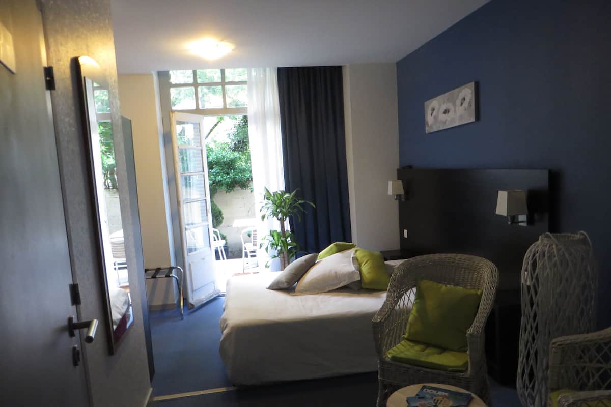 A hotel room with a bed, a wicker chair, a small table with magazines, a nightstand, and a view of the outside.