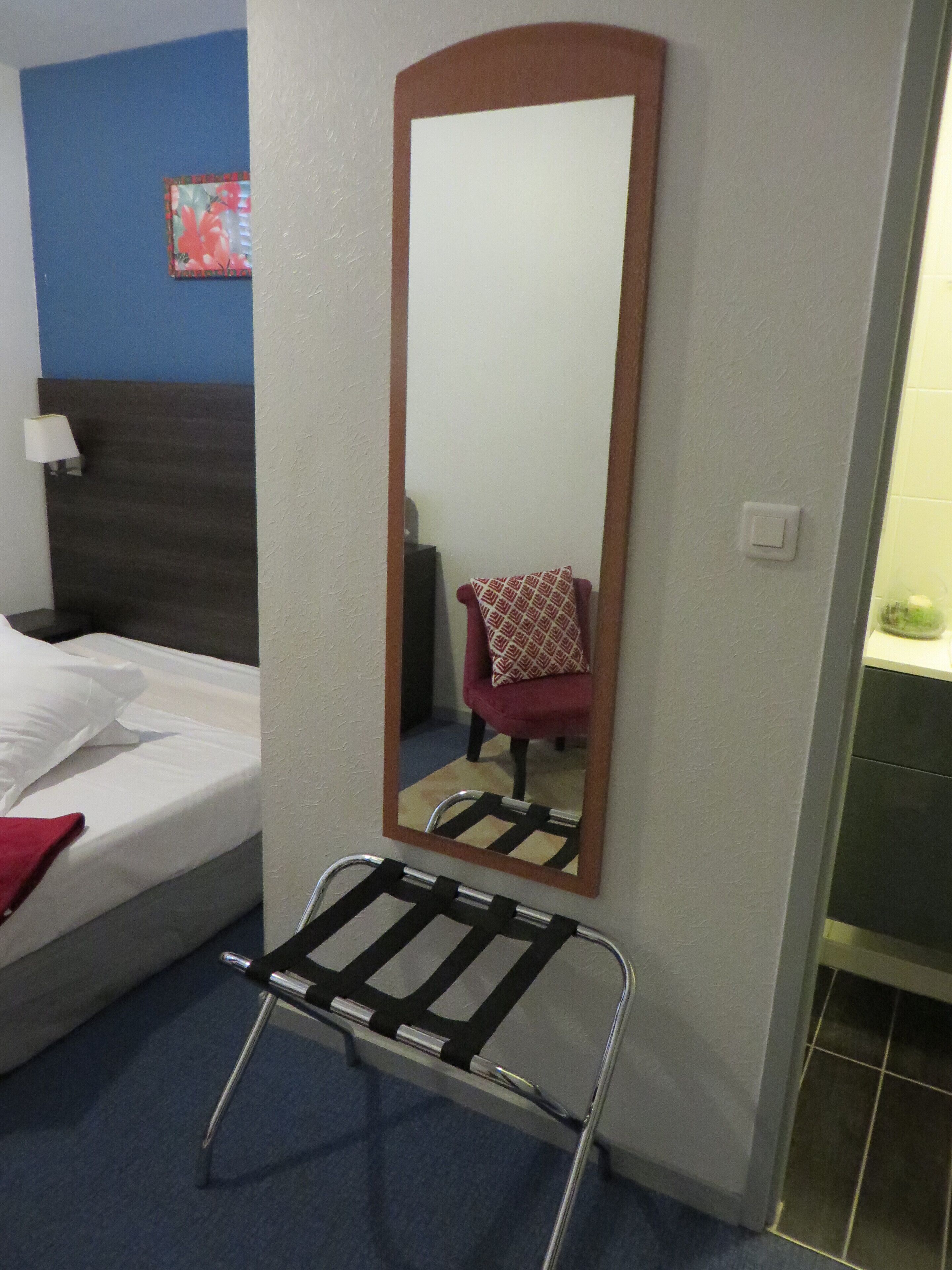 A hotel room with a bed, a mirror, a chair, and a metal rack.