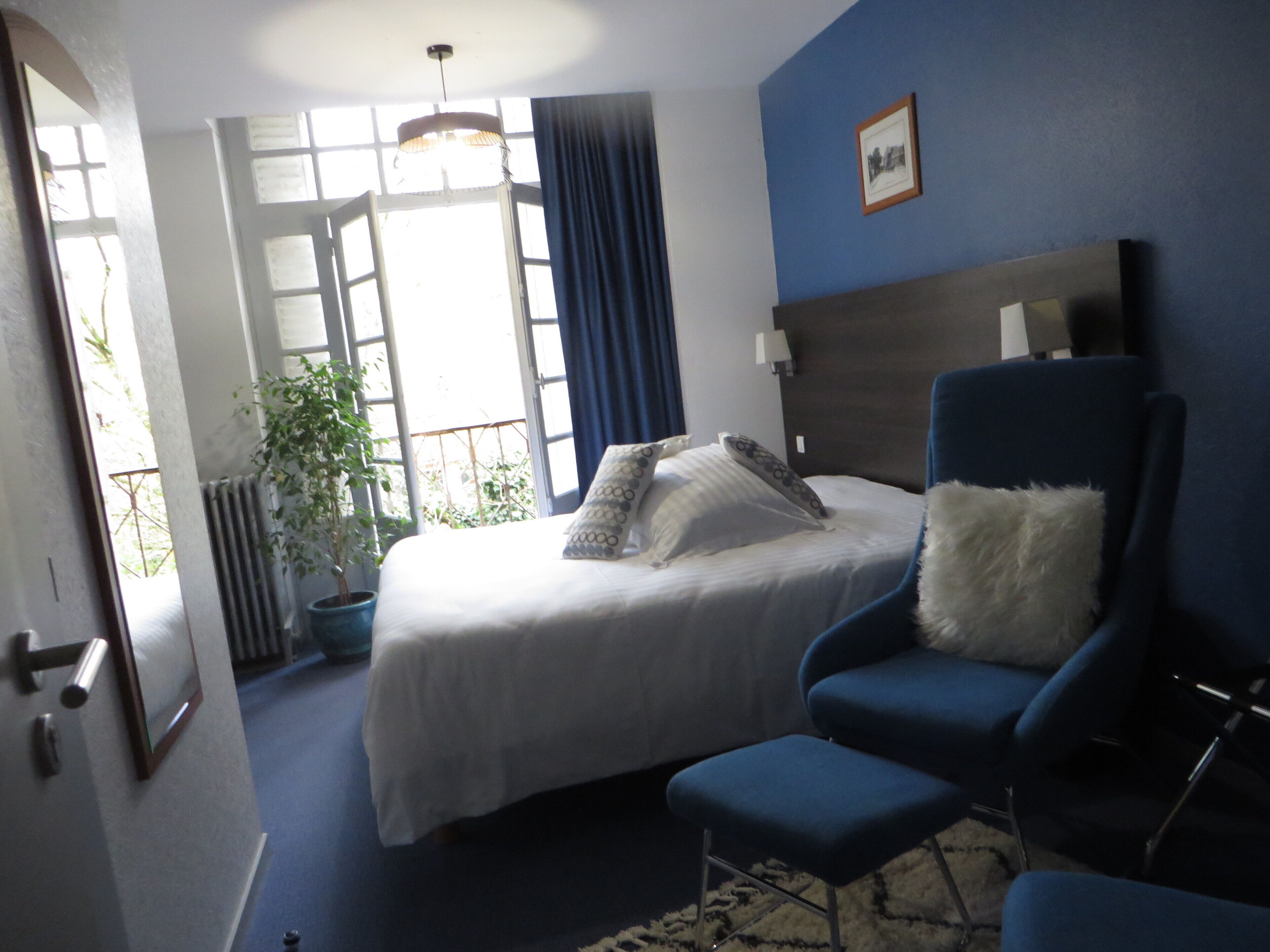 Ein Hotelzimmer mit einem Bett, einem blauen Sessel, einem weißen Kopfkissen, einem blauen Vorhang und einem gerahmten Bild an der Wand.
