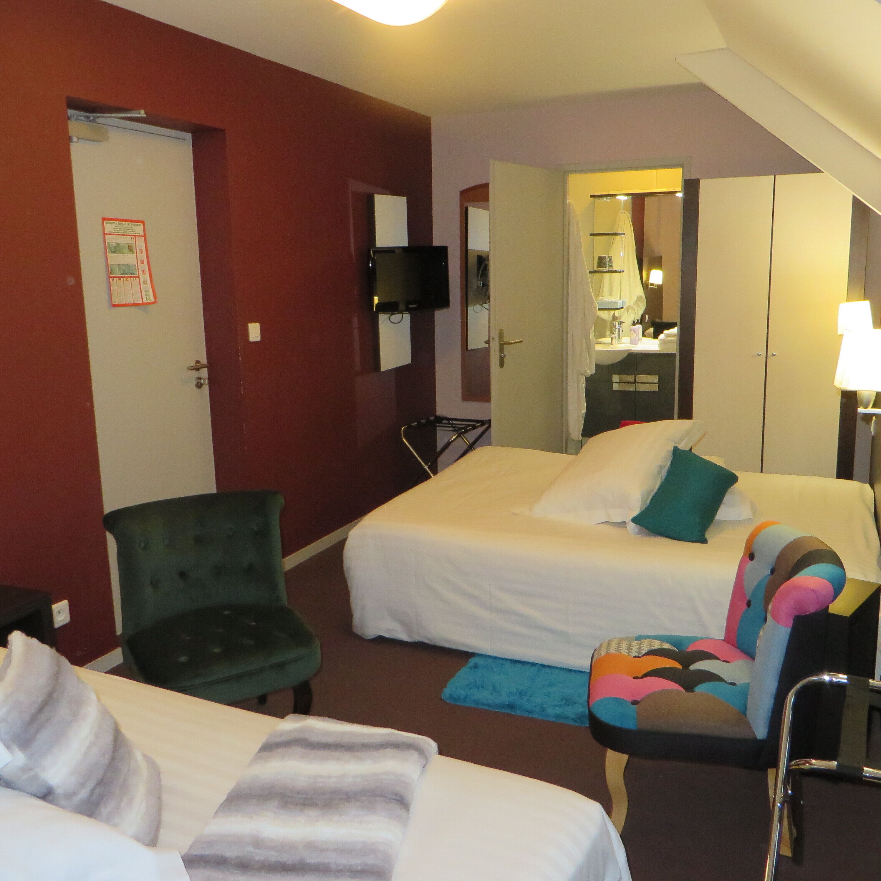 Hotelkamer met twee bedden, een groene fauteuil, een kleurrijke stoel, een bureau met lamp, en een badkamer zichtbaar door een geopende deur.