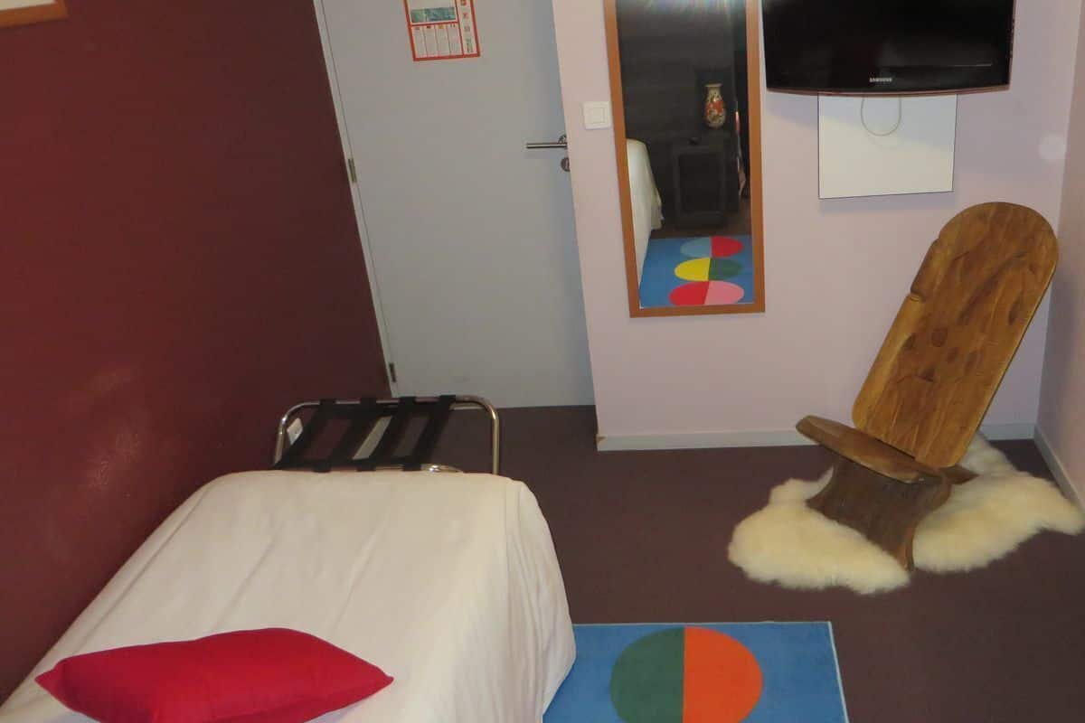 A hotel room with a bed, a chair with a fur seat, a mirror, and a television on the wall.