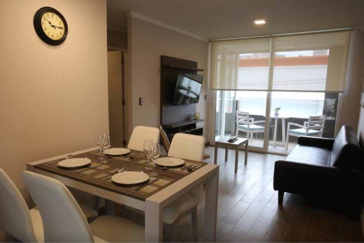A dining area with a table set for four, a flat-screen TV, and a view of the sea through large windows.