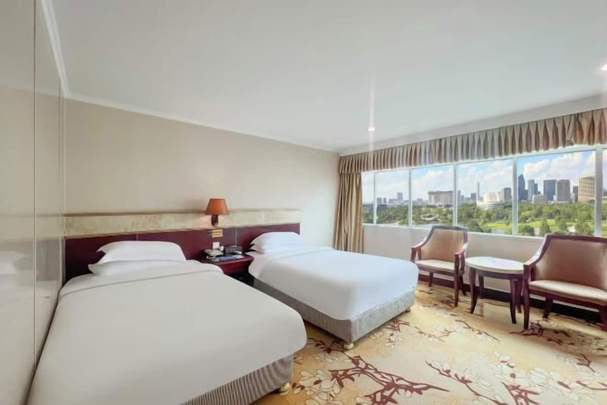 Habitación de hotel con dos camas, una mesita de noche, una lámpara, una silla, una pequeña mesa y un amplio ventanal con vistas a la ciudad.