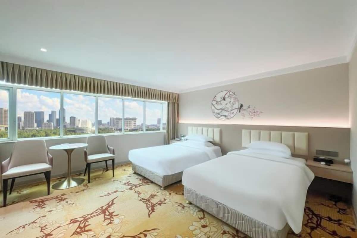 Habitación de hotel con dos camas, una mesita pequeña, dos sillas, un ventanal grande con vista a la ciudad y un adorno de pared.
