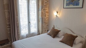 A bedroom with a large bed, stone wall, window with curtains, and a painting on the wall.