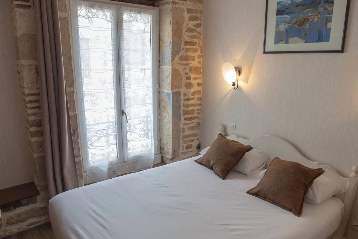 Une chambre avec un grand lit, un mur en pierre, une fenêtre avec des rideaux et un tableau accroché au mur.