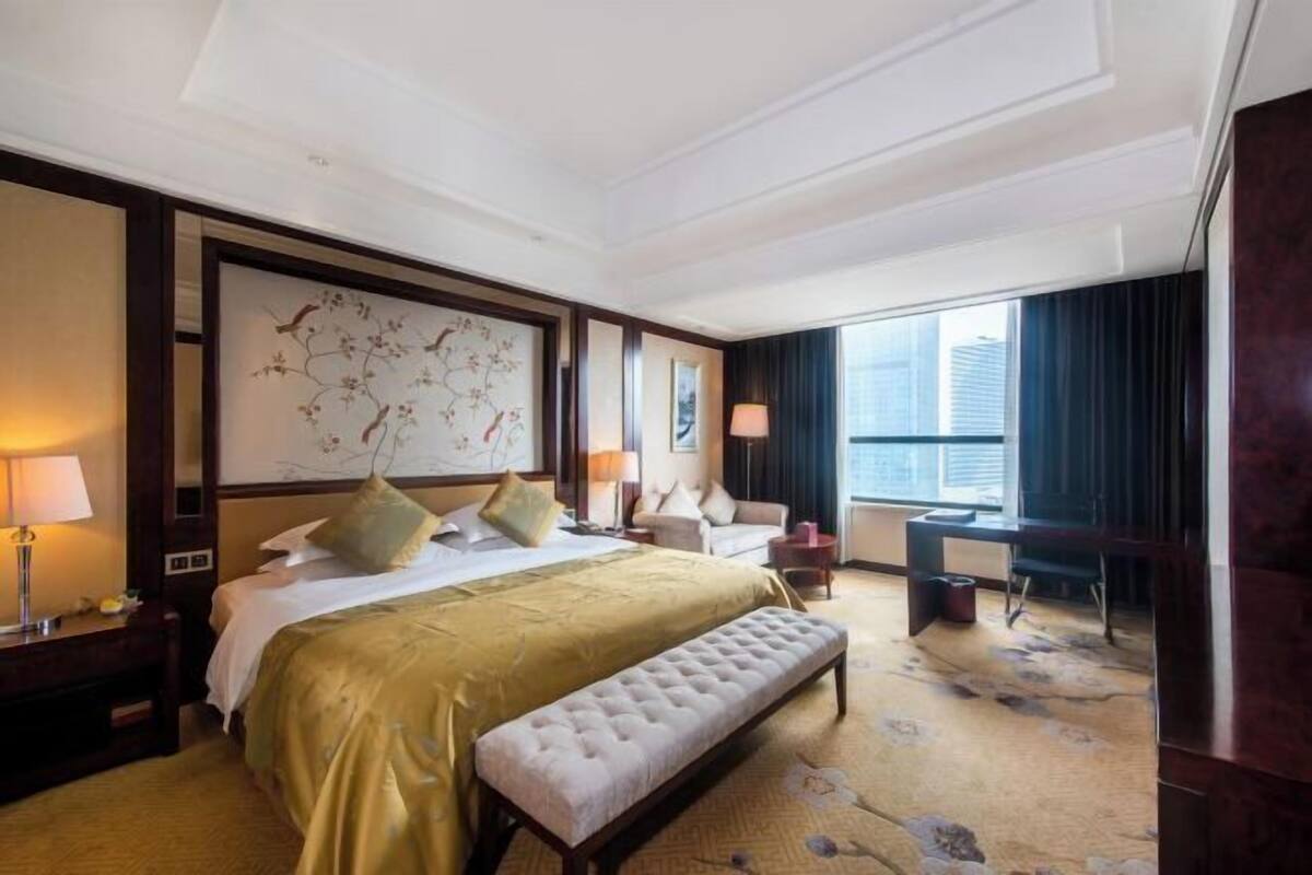 A hotel room with a large bed, a bench, a desk, and a view of the city.