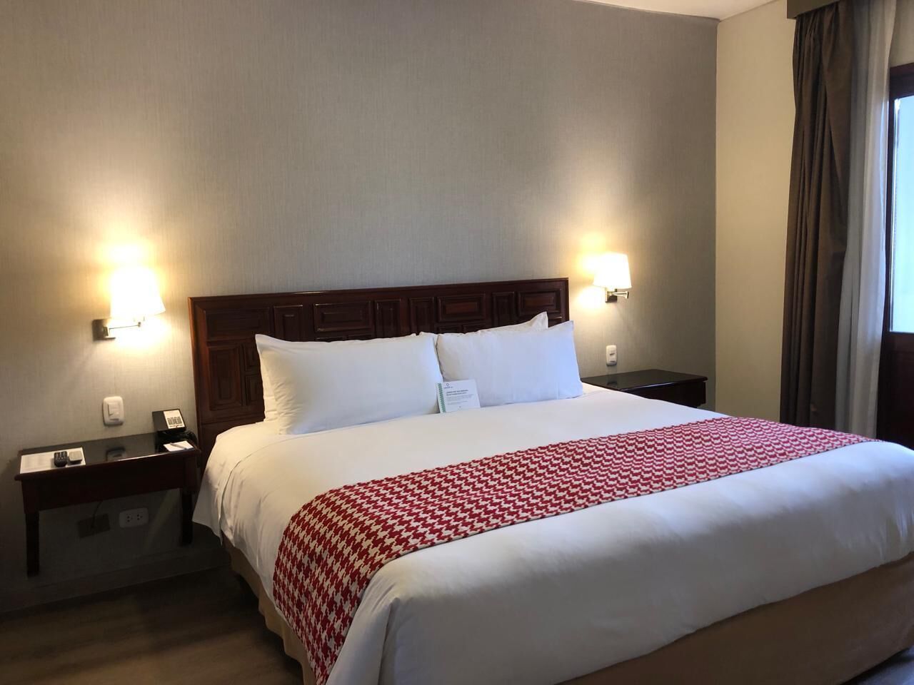 Habitación de hotel con una cama grande, dos mesitas de noche, cabecera de madera y dos lámparas de pared.