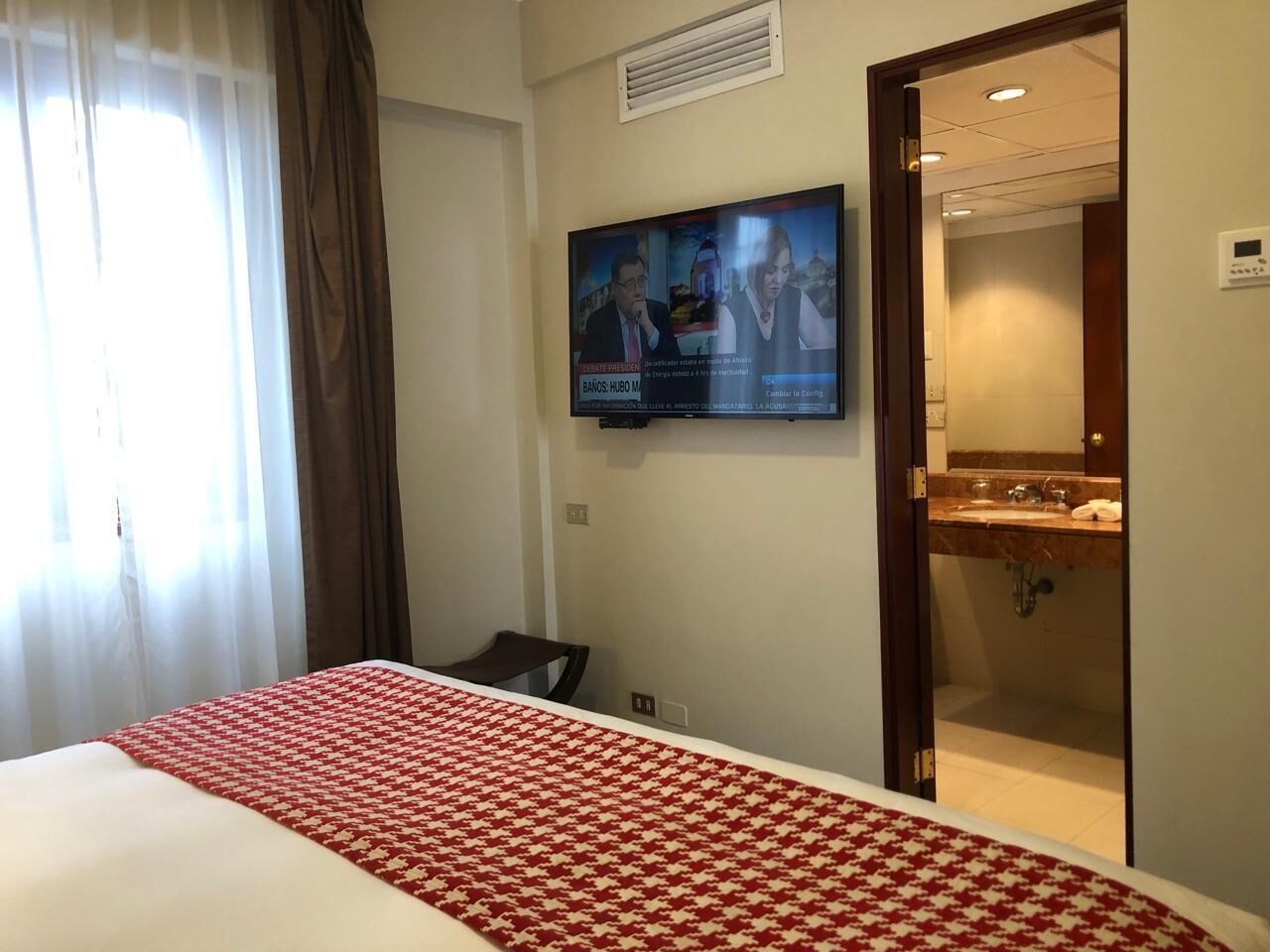 Une chambre d’hôtel avec un lit, un téléviseur fixé au mur et une salle de bain visible à travers une porte ouverte.