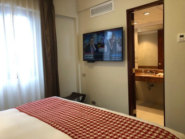 Habitación de hotel con cama, televisor montado en la pared y baño visible a través de una puerta abierta.