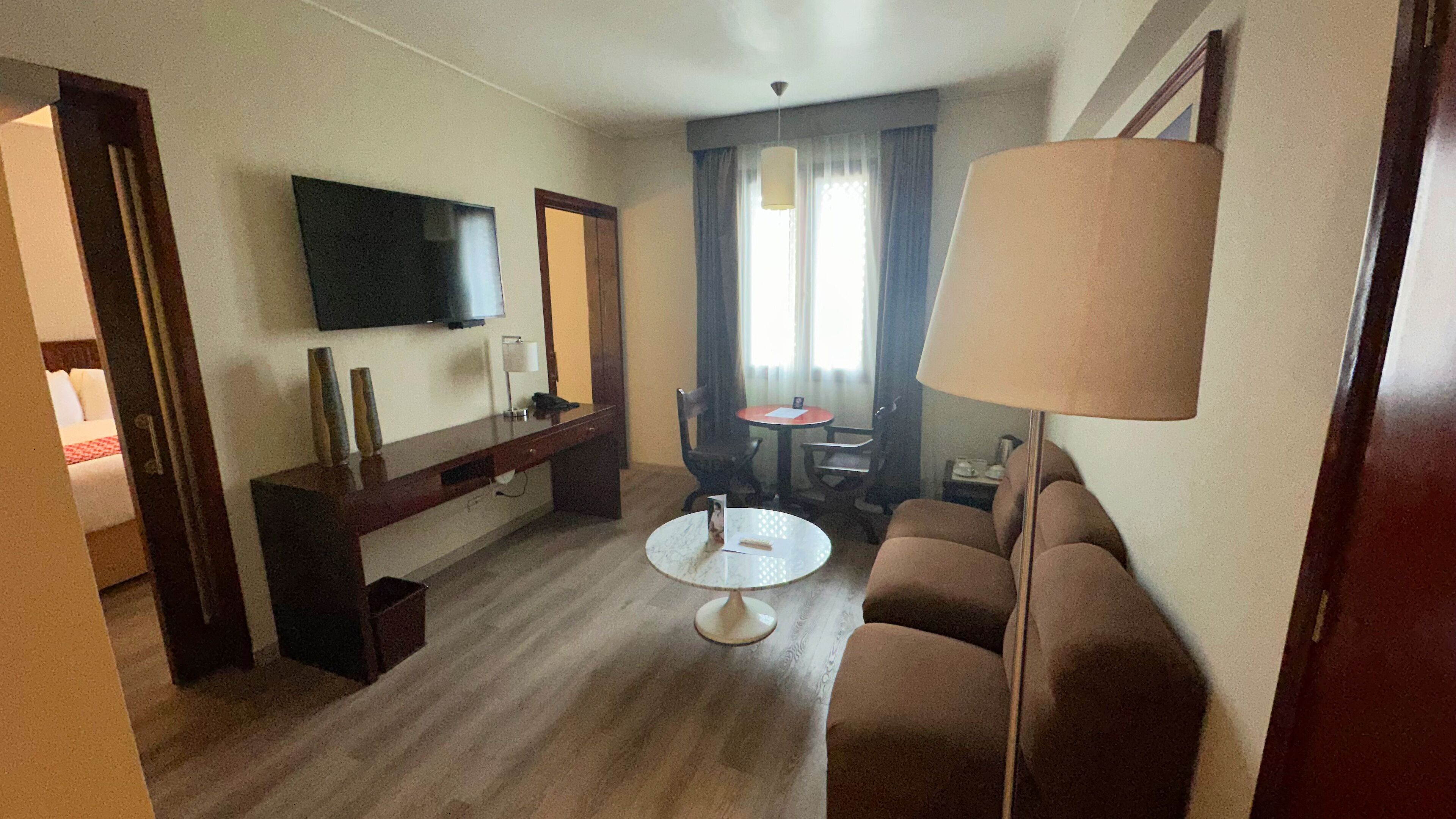 A hotel room with a sofa, a small round table, a TV mounted on the wall, a desk with a chair, and a window with curtains.