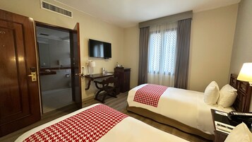 A hotel room with two beds, a desk, a chair, a TV, and a window with curtains.