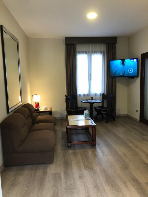 Una sala de estar con un sofá marrón, una mesa de centro, un televisor montado en la pared y un ventanal con cortinas.