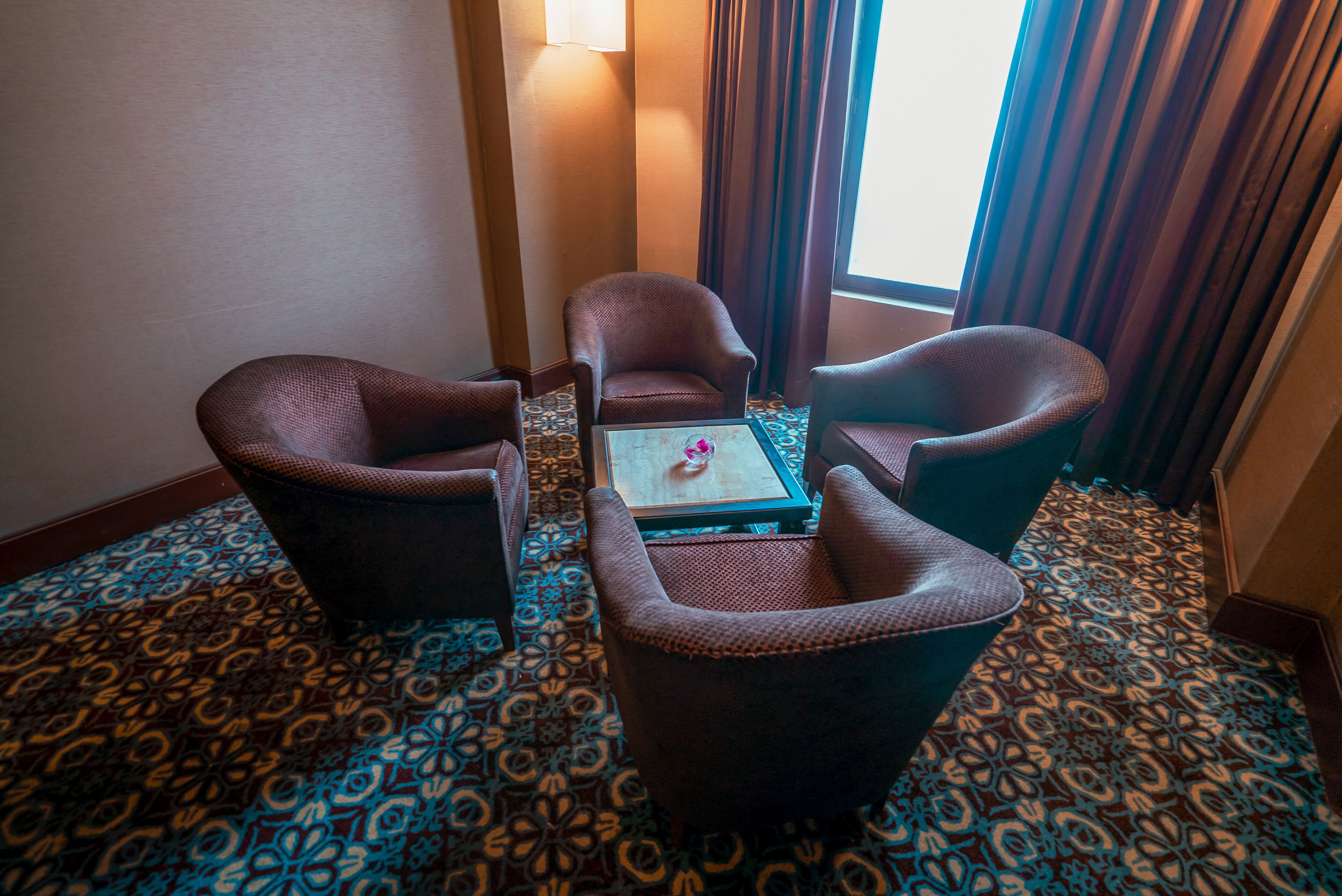 A hotel room with a patterned carpet, a small table with a vase, and two armchairs.