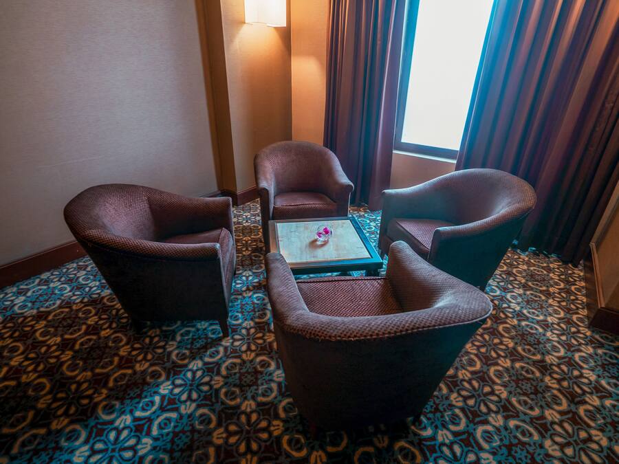 A hotel room with a patterned carpet, a small table with a vase, and two armchairs.
