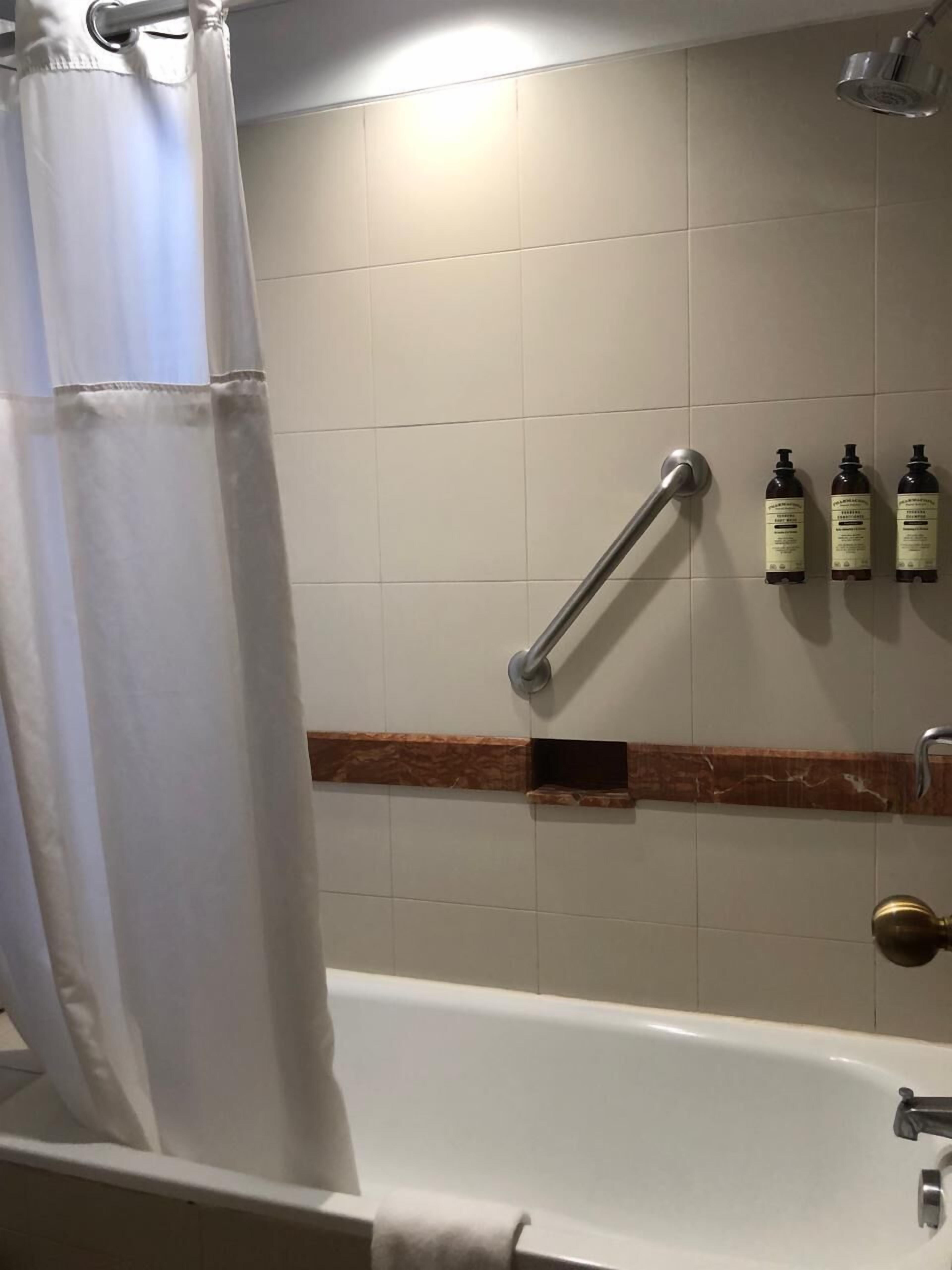 A bathroom with a bathtub, a shower curtain, a grab bar, and three bottles of shampoo on a shelf.