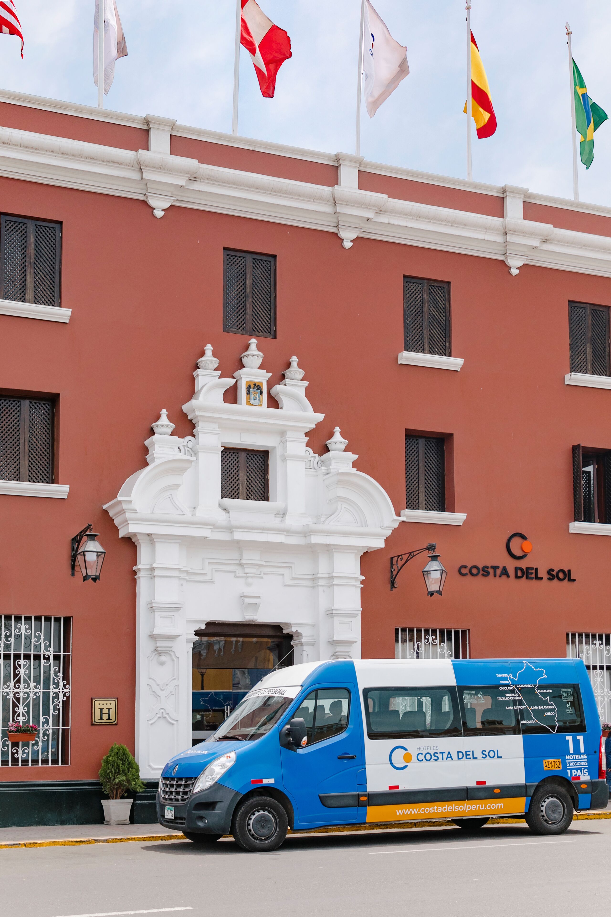 A building with a white facade and a blue van parked in front. Flags are visible above the entrance.