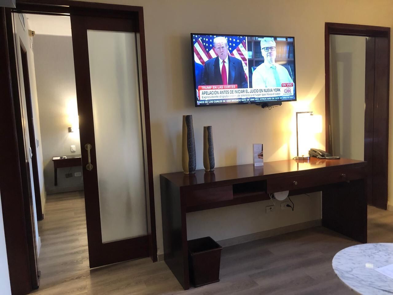 Habitación de hotel con televisión montada en la pared mostrando noticias, una mesita de consola de madera con jarrones decorativos, una lámpara de noche y un pequeño cubo de basura.