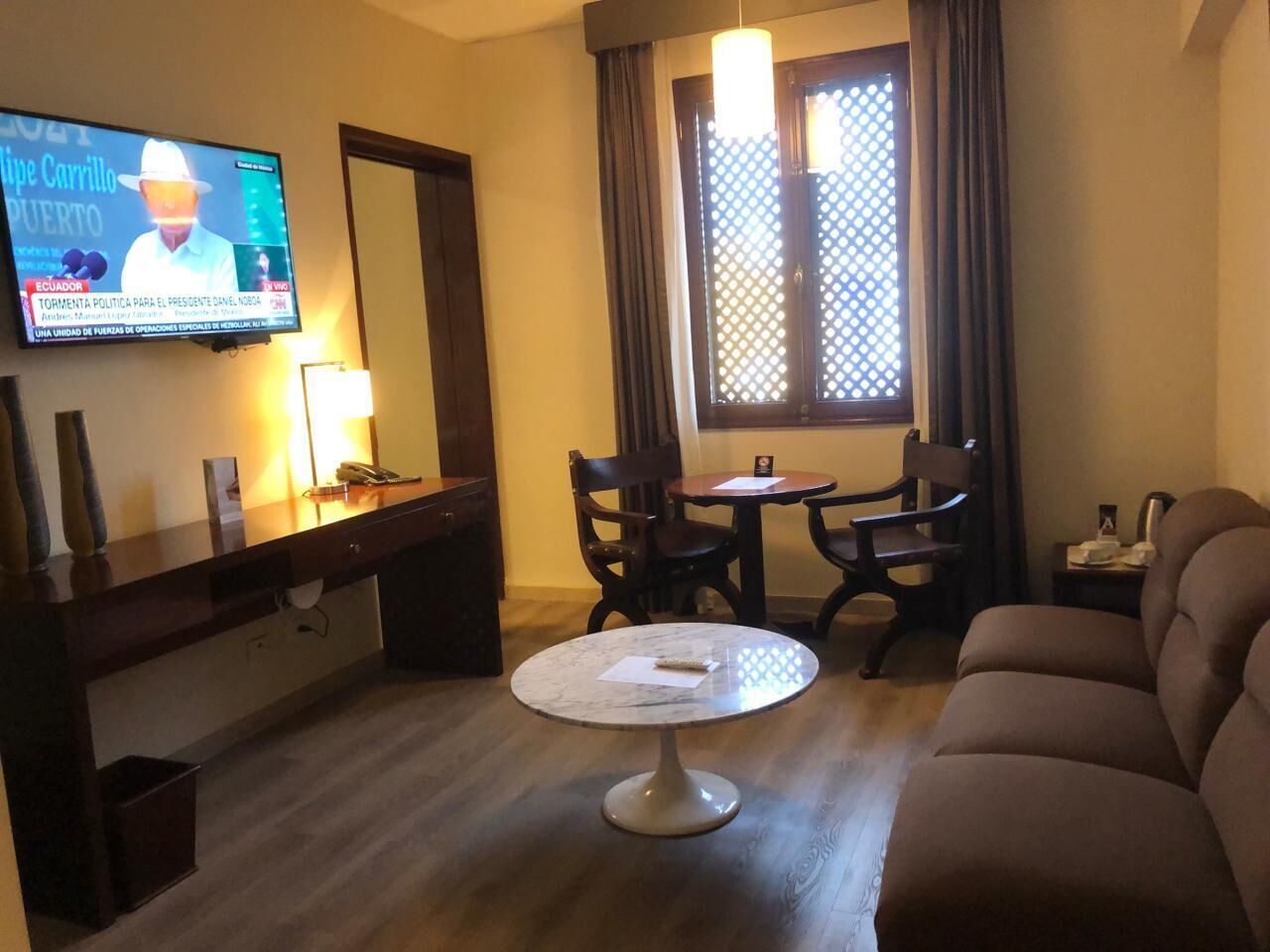 Habitación de hotel con una televisión mostrando un noticiero, una mesa redonda con sillas, un sofá y una ventana con cortinas.