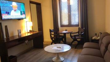 Habitación de hotel con una televisión mostrando un noticiero, una mesa redonda con sillas, un sofá y una ventana con cortinas.