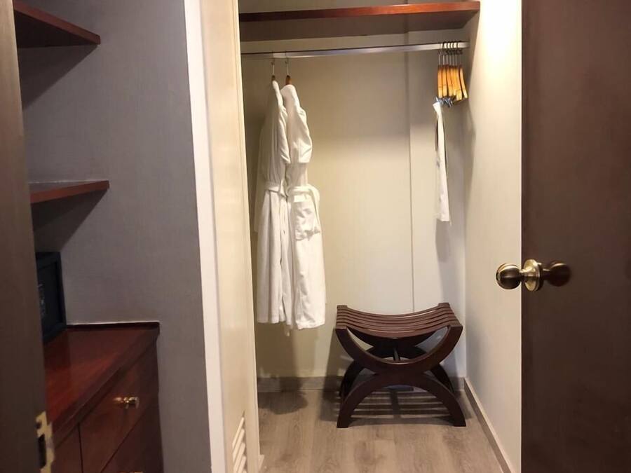 A hotel room with a wooden wardrobe, a bench, and a walk-in closet with hanging robes.