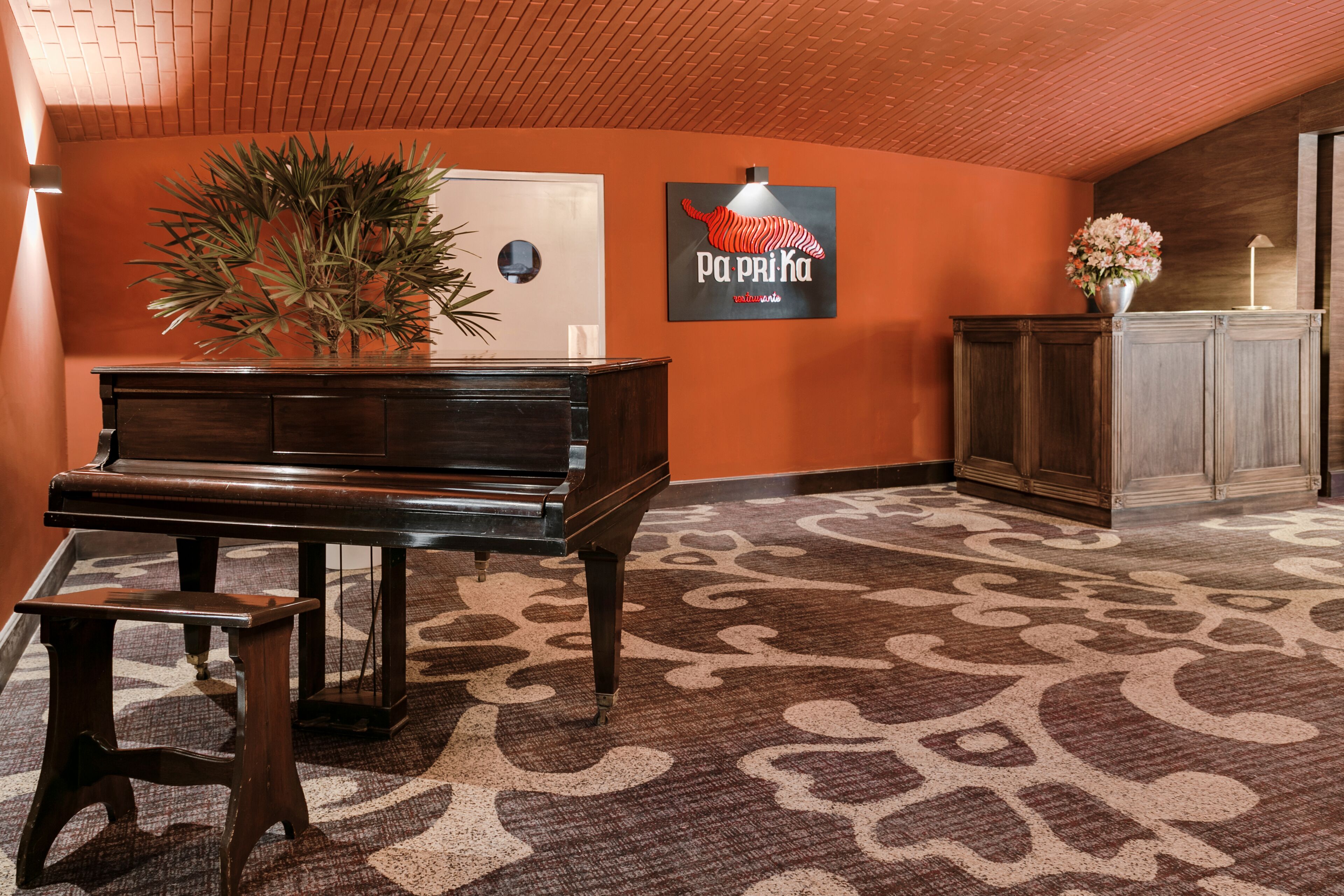 Un piano de cola en un vestíbulo con una alfombra estampada, una planta en maceta y un cartel en la pared.