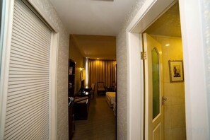 Un couloir d’hôtel avec une porte fermée sur la gauche, un bureau avec une chaise et une lampe au centre, et un lit avec une tête de lit sur la droite.